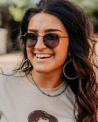 Woman wearing sunglasses and a gray t-shirt with a logo, outdoors.