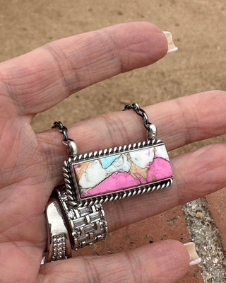 Hand holding a small silver pendant with colorful stone inlay against a brown background