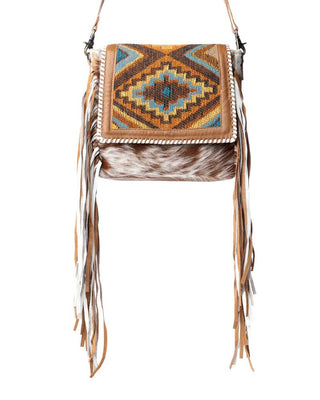 A western-style crossbody bag featuring a geometric patterned flap in brown, orange, and blue, a brown faux fur body, and long tan leather fringe on the sides, shown against a plain white background.