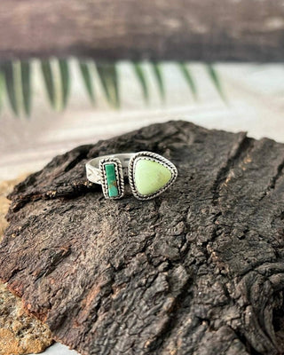 A silver ring with a large light green teardrop-shaped stone and a smaller green rectangular stone, displayed on a textured piece of dark bark.