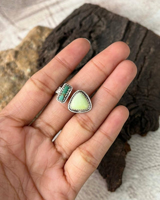 Hand wearing two rings with green and gemstones on a textured surface