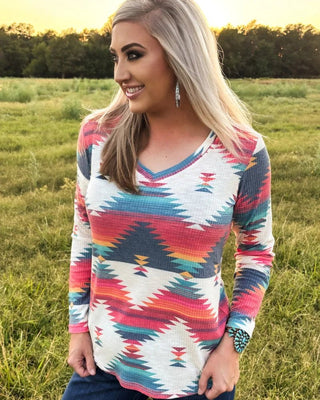 A woman wearing a long-sleeve waffle-knit V-neck top with a colorful Southwestern geometric pattern, standing in a grassy field at sunset.