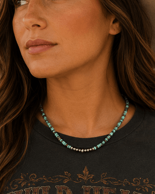 A woman wearing a beige cowboy hat and a dark gray graphic t-shirt with a longhorn skull and 'Wild Heart' text, accessorized with a turquoise beaded necklace, set against a rustic wooden wall.