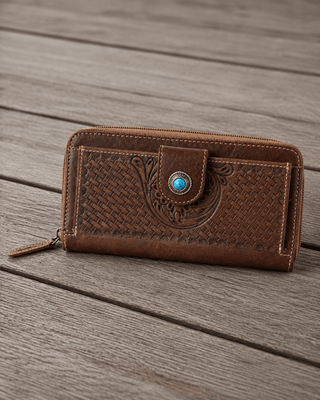 A brown leather zip-around wallet with embossed western-style floral and basketweave patterns, featuring a snap-button closure with a turquoise accent, resting on a weathered wooden plank surface.
