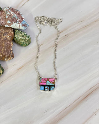 Necklace with a square pendant featuring colorful stones on a wooden surface with rocks in the background.