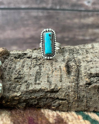 A silver ring with a rectangular turquoise stone, displayed on a textured piece of wood.