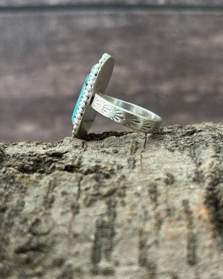 A sterling silver ring with a turquoise stone and small clear accent stones, shown resting on a textured piece of wood with a blurred natural background.