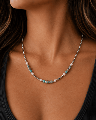 A close-up of a woman wearing a delicate beaded necklace with turquoise, white, and gray beads over a black ribbed tank top.