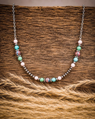 A beaded necklace with silver-toned chain and beads, featuring turquoise, pink, white, and patterned accent beads, displayed on a rustic wood surface with dried grass in the foreground.