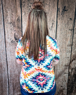 A woman viewed from behind wearing a short-sleeve white t-shirt with a vibrant multicolor geometric tribal pattern, standing against a rustic wooden plank wall.