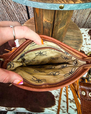 A close-up of a brown leather crossbody bag being held open by a hand, showing the tan interior lined with black logos. The background features a rustic wooden barrel and a cowhide rug.