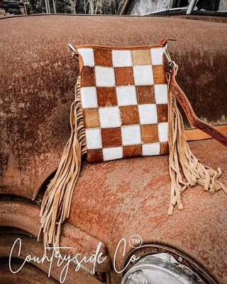 A western-style checkered clutch bag with fringe tassels, displayed on the rusted hood of a vintage car. The bag features a brown and white checkerboard pattern made from cowhide patches, with a brown leather strap and long tan fringe.