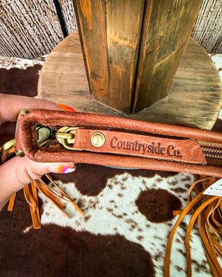 A close-up of a person's hand holding a brown leather zippered pouch with a brass zipper pull and a leather tab engraved with 'Countryside Co.' The background features a rustic wooden pole and a brown and white cowhide rug.