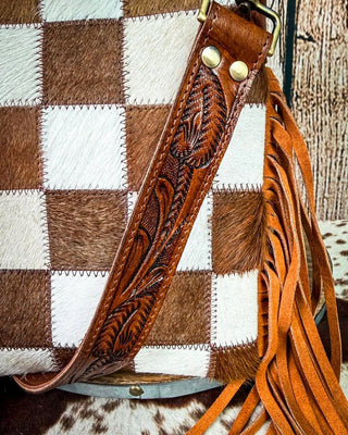 Close-up of a western-style handbag with a brown and white checkered hide pattern, a tooled leather shoulder strap, and long orange-brown fringe tassels.