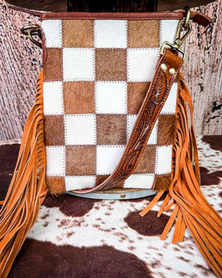 A western-style bucket bag featuring a brown and white checkerboard pattern on the front, adorned with long orange fringe on the sides and a tooled brown leather strap, displayed on a cowhide surface.