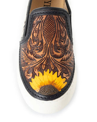A single slip-on shoe with a black leather upper featuring intricate brown floral tooling and a bright yellow sunflower design on the toe, finished with a thick white rubber sole.