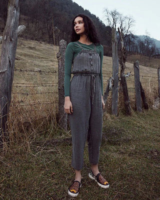 A woman stands outdoors in a rural landscape wearing a dark gray sleeveless jumpsuit over a green long-sleeve top, with a rustic fence and hills in the background.
