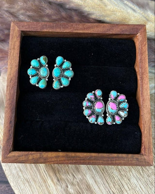 A pair of turquoise and pink stud earrings displayed on a black velvet insert inside a wooden box, set against a rustic wood and faux fur background.