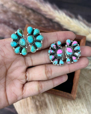 A pair of silver-toned stud earrings with turquoise and pink stones, held in a person's hand. The earrings feature a cluster of teardrop-shaped turquoise stones and smaller pink stones set in a textured silver bezel.