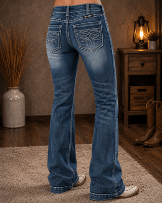 A person shown from the back wearing blue western-style flare jeans with decorative stitching on the back pockets, standing on a rug in a rustic indoor setting with a lamp, basket, and boots nearby.
