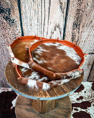 A rustic leather and cowhide tray or basket with a brown and white spotted hide pattern and reddish-brown leather trim, displayed on a wooden stand against a wood plank backdrop.