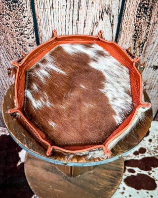 A round cowhide tray with a brown and white pattern, bordered by a rust-orange leather trim, displayed on a rustic wooden stand against a wood plank background.