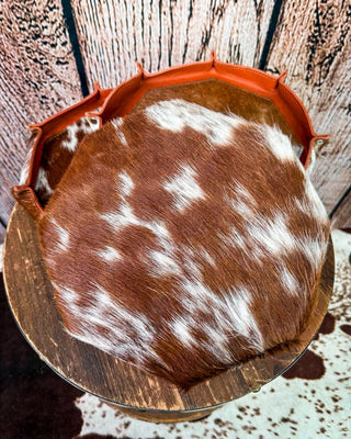 A round cowhide tray with a brown and white pattern, bordered by a rust-orange leather trim, displayed on a rustic wooden stand against a wood plank background.