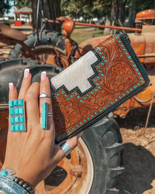 Hand holding a tooled leather wallet with turquoise accents in front of a tractor.