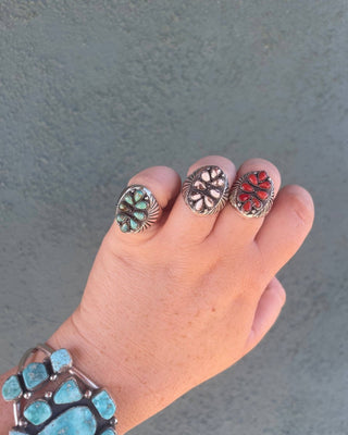 A hand wearing three silver-toned rings with inlay stones in green, white, and red, along with a matching turquoise bracelet, set against a textured gray background.