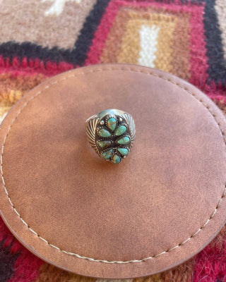 A silver-toned ring with a floral cluster of turquoise stones, displayed on a round brown leather pad over a colorful woven rug.
