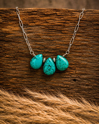 A silver chain necklace with three teardrop-shaped turquoise stones, displayed on a rustic wooden surface with dried grass in the foreground.