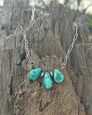 A silver chain necklace with three turquoise-colored stone pendants, displayed on a textured piece of weathered wood outdoors.