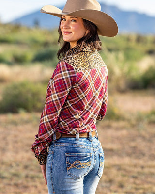 A woman wearing a red and beige plaid western shirt with a leopard-print collar, blue jeans, and a tan cowboy hat, standing in an outdoor desert landscape.