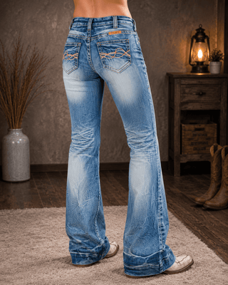 A person shown from the waist down wearing light blue flared denim jeans with a faded wash and decorative stitching on the back pockets, standing in a rustic indoor setting with boots and a lamp in the background.