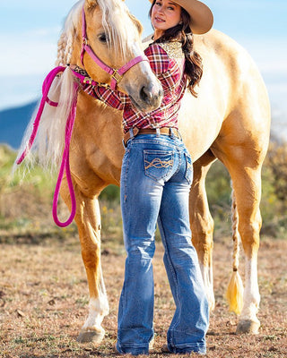 A woman in a cowboy hat, red plaid shirt, and blue jeans stands beside a light tan horse outdoors, holding its reins. The horse has a bright pink halter and matching pink lead rope.