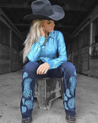 A woman wearing a black cowboy hat, a shiny light blue button-up shirt, dark blue jeans with light blue paisley embroidery, and boots, sitting on a wooden block in a rustic barn setting.