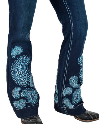 Close-up of dark blue denim jeans with large light blue paisley embroidery on the lower legs and white contrast stitching, shown on a model's legs with black boots visible at the bottom.