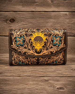 A tooled leather clutch wallet with a prominent yellow sunflower design and intricate brown floral scrollwork, accented with turquoise blue details, displayed on a rustic wooden surface.