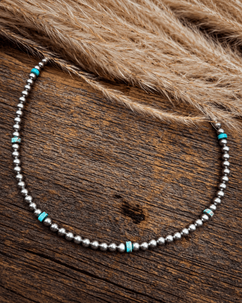A beaded necklace with silver-toned round beads and small turquoise-colored spacer accents, displayed on a rustic wooden surface with dried grass in the background.