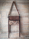 Front view of the Sandy Rosewood Hand-Tooled Bag hanging on a brick wall. 